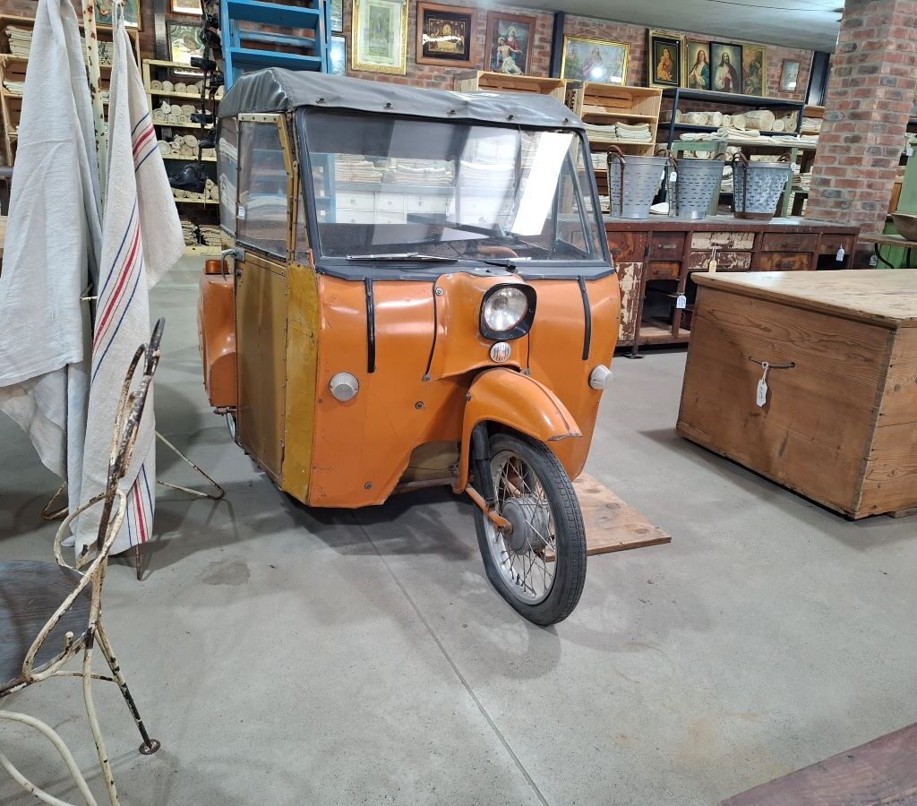 A quirky and humorous photo taken during the Wild Buggers drive showing a strange, vintage three-wheeled vehicle—an unconventional classic car—on display at the Kooperasie Stories antique store in Simondium.