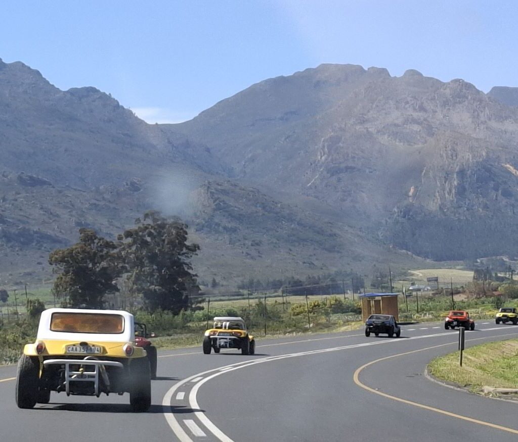 A close-up action shot of a convoy of Wild Buggers Beach Buggies driving on a scenic road, captured from a vehicle behind the group, emphasizing the movement, speed, and fun of the club's VW air-cooled vehicles.