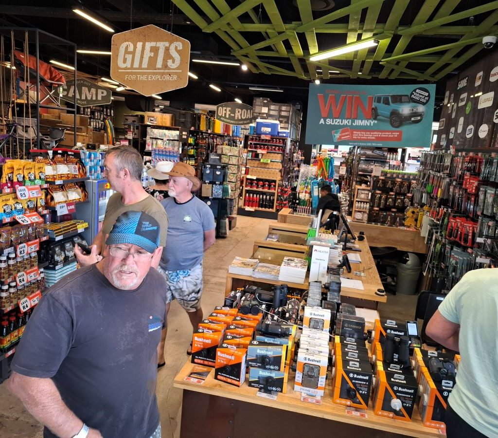 A photo showing several members of the Wild Buggers Beach Buggy Club—including Anthony, Brent, Rhoodie, Chris, Anton, and Jacques—browsing and laughing inside an outdoor camping supply store in Paarl during a stop on their scenic drive.