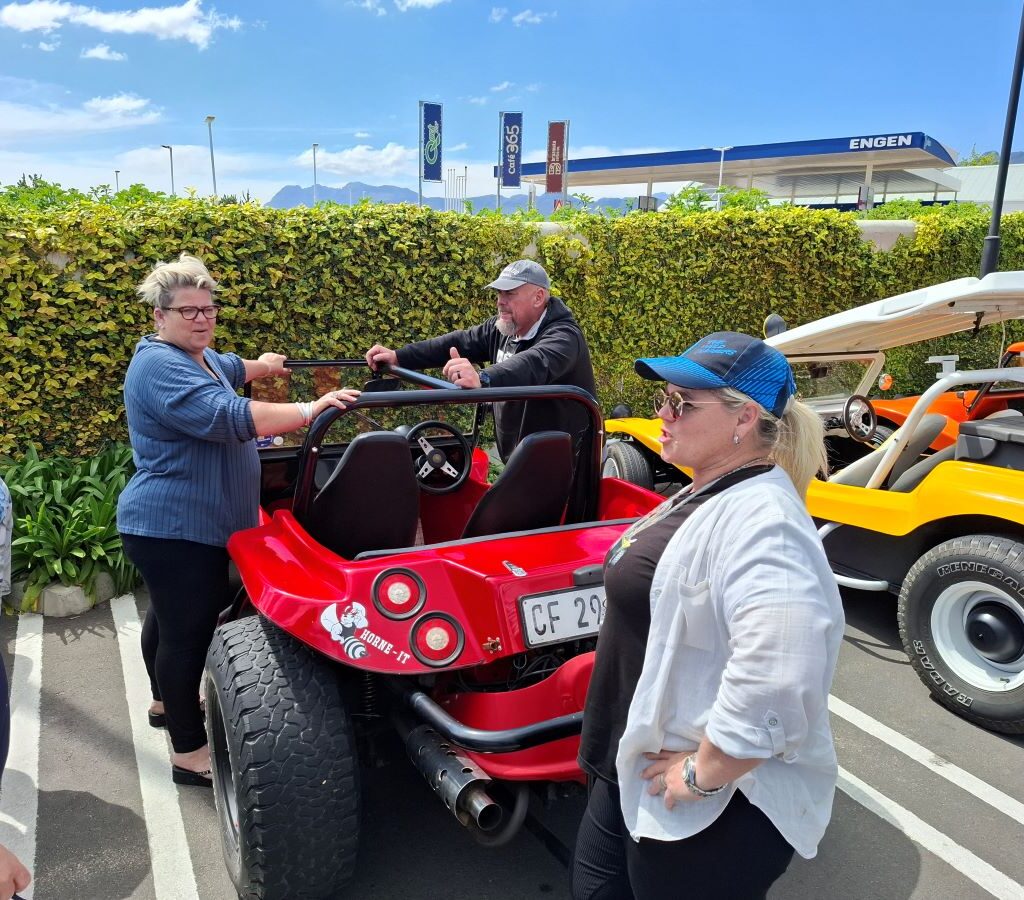 A cheerful and candid photo of a group of Wild Buggers members laughing and socializing next to their colorful VW air-cooled vehicles in the parking area of the Paarl shopping centre, waiting to restart their scenic drive.