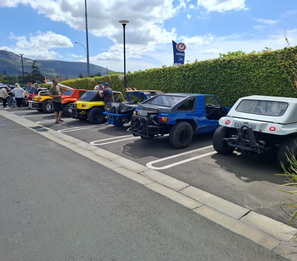 A relaxed, wide shot of the Wild Buggers members grouped together in the Paarl shopping centre parking lot, laughing and enjoying a chat while keeping a casual eye on their line of parked beach buggies.