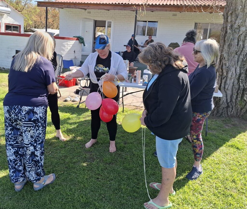 A funny photo of the Wild Buggers "Ladybugs" (Sandra, Sam, Michelle, Mickey, Yolande, and Linda) gathered at the Bergriver Resort camp campsite after the morning drive, intensely focused on blowing up and tying balloons for a mysterious club activity.