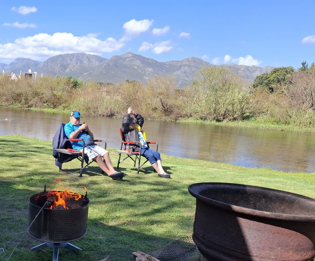A warm, peaceful photo of Thys and his wife relaxing in camping chairs by the river at the Bergriver Resort camp, with a small campfire burning cozily beside them in the early evening. The scene captures the tranquility and natural beauty of the campsite.