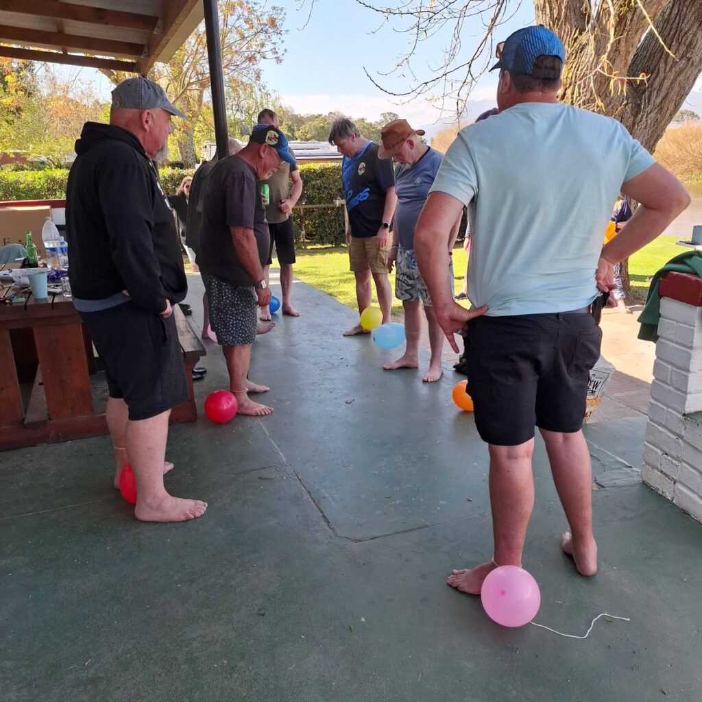 A hilarious action photo at the Bergriver Resort camp showing the Wild Buggers men—including Anthony, Brent, Rhoodie, Chris, Anton, and Jacques—standing awkwardly with balloons tied to their legs, preparing to start a chaotic balloon-popping game against each other.