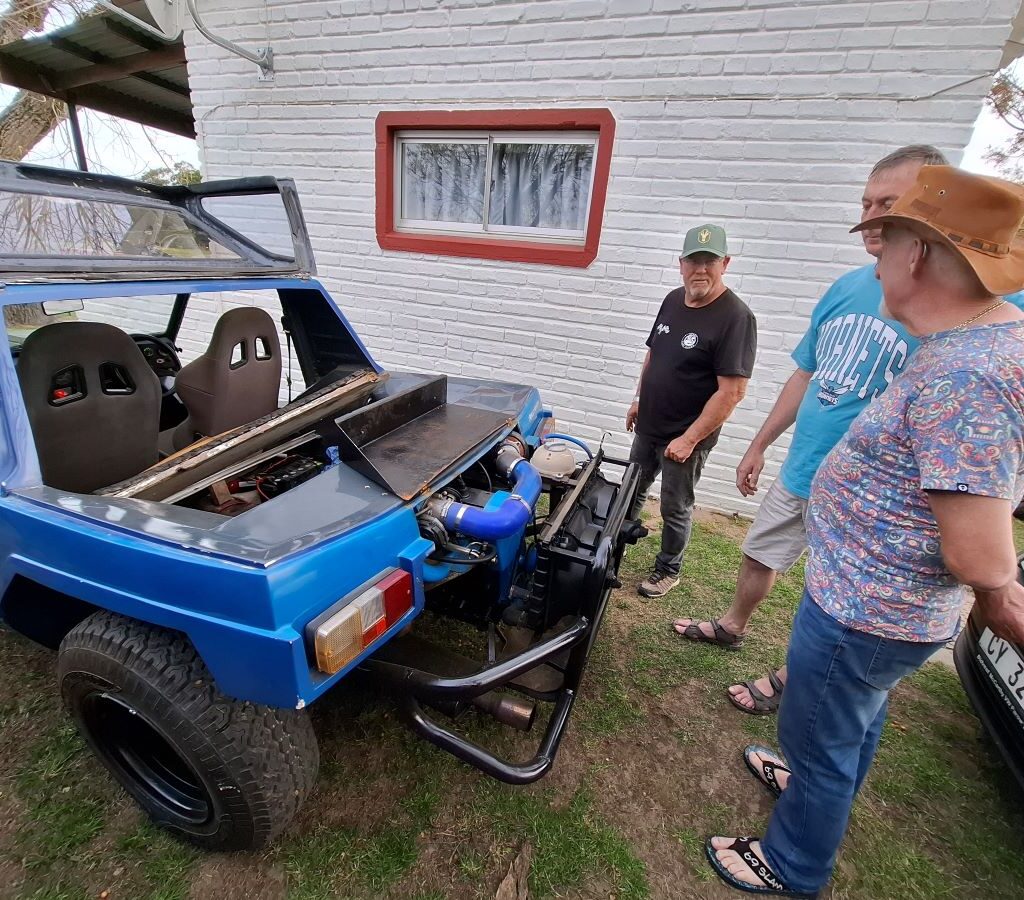 A candid and funny photo from the Bergriver Resort camp showing two Wild Buggers, Anthony and Jacques, leaning over the engine bay of Rhoodie’s beach buggy, which has a non-traditional water-cooled Golf engine conversion installed. The scene captures the interest and debate over VW modifications.