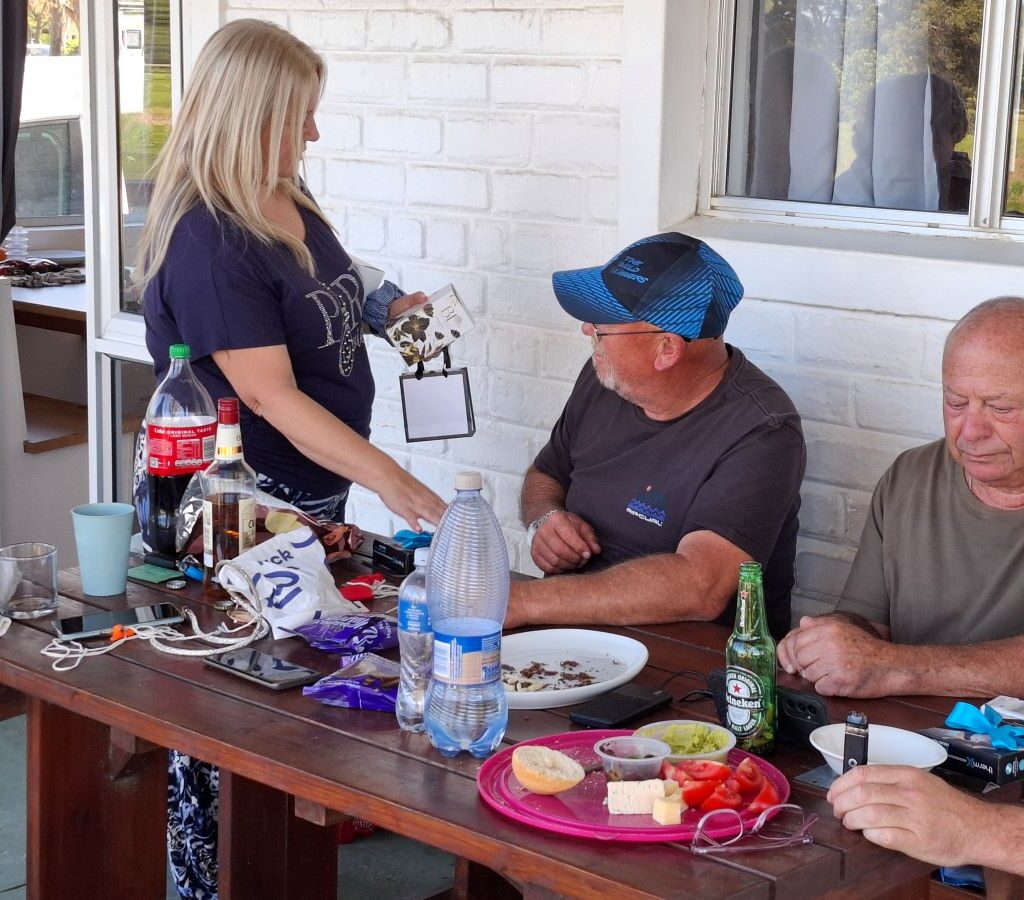 A joyful photo taken at the Bergriver Resort camp showing Sam Van Der Weijde presenting a prize to Anthony Jenkins, who is smiling broadly, celebrating his win in the chaotic balloon-popping competition. The scene captures the fun and camaraderie of the Wild Buggers at the kuierspot.