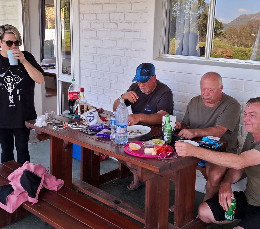 A candid photo of four Wild Buggers members—Yolande, Anthony, Clive, and Rhoodie—seated together and engaged in a casual, friendly chat around a table at the Bergriver Resort camp campsite.
