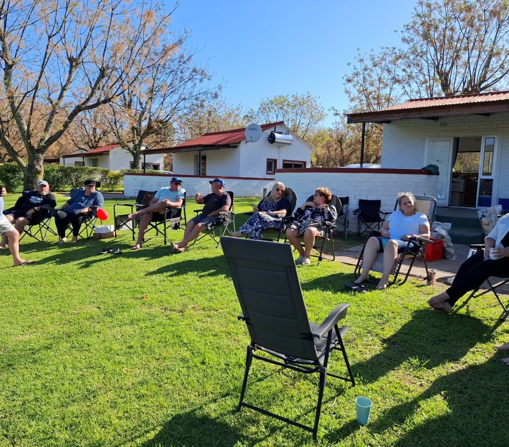 A wide, candid photo showing multiple members of the Wild Buggers Beach Buggy Club sitting in their camping chairs on the lawn at the Bergriver Resort camp, completely relaxed and casually chatting, highlighting the weekend's peaceful, social atmosphere.