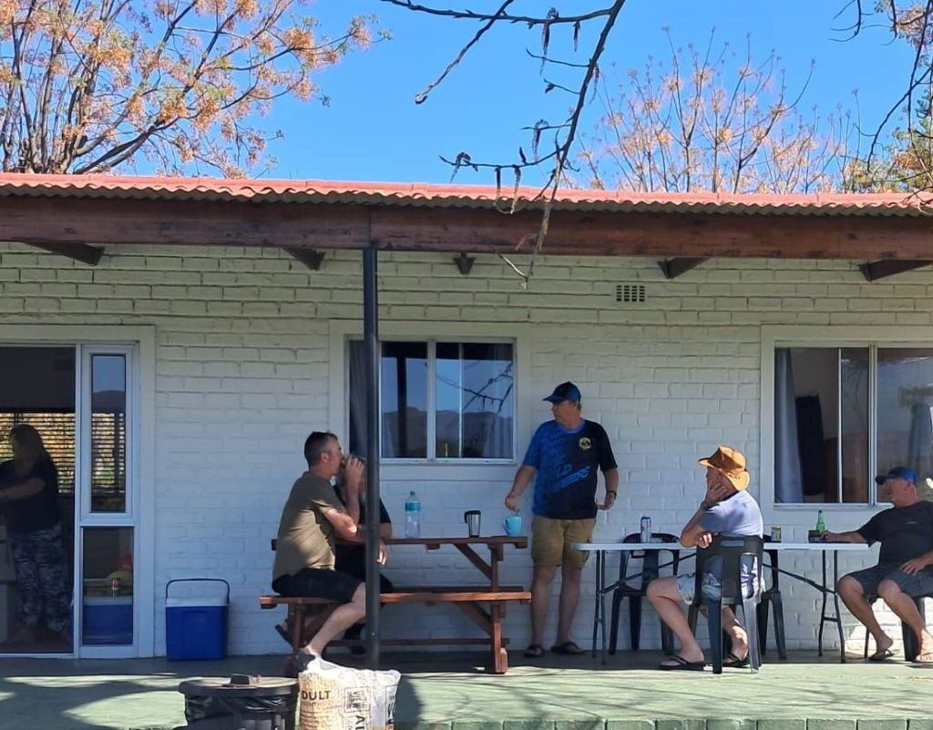 A warm, candid photo of several Wild Buggers members seated at a table on the stoep (porch) of a building at the Bergriver Resort camp, engaged in a relaxed conversation in the late afternoon sun.