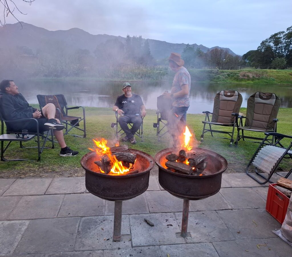 A warm, inviting photo of three Wild Buggers members—Anthony, Brent, and Jacques—relaxing and chatting around a glowing campfire in the early evening at the Bergriver Resort camp. The scene evokes camaraderie and the relaxed atmosphere of a club gathering.