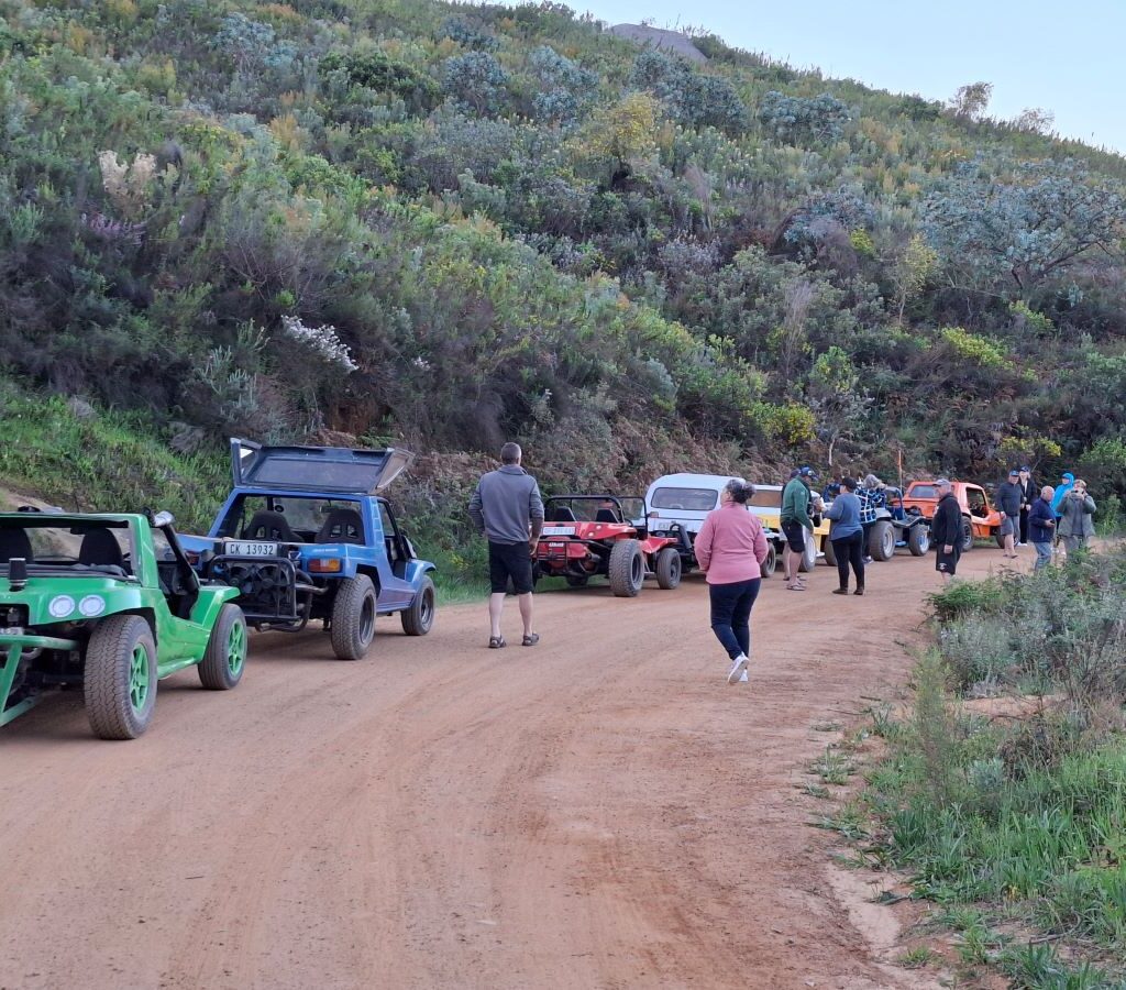 A spectacular photo of the Wild Buggers members gathered on the Paarlberg mountain, enjoying a beautiful sundowner with their beach buggies parked nearby, overlooking a scenic view in the warm, late-afternoon light.