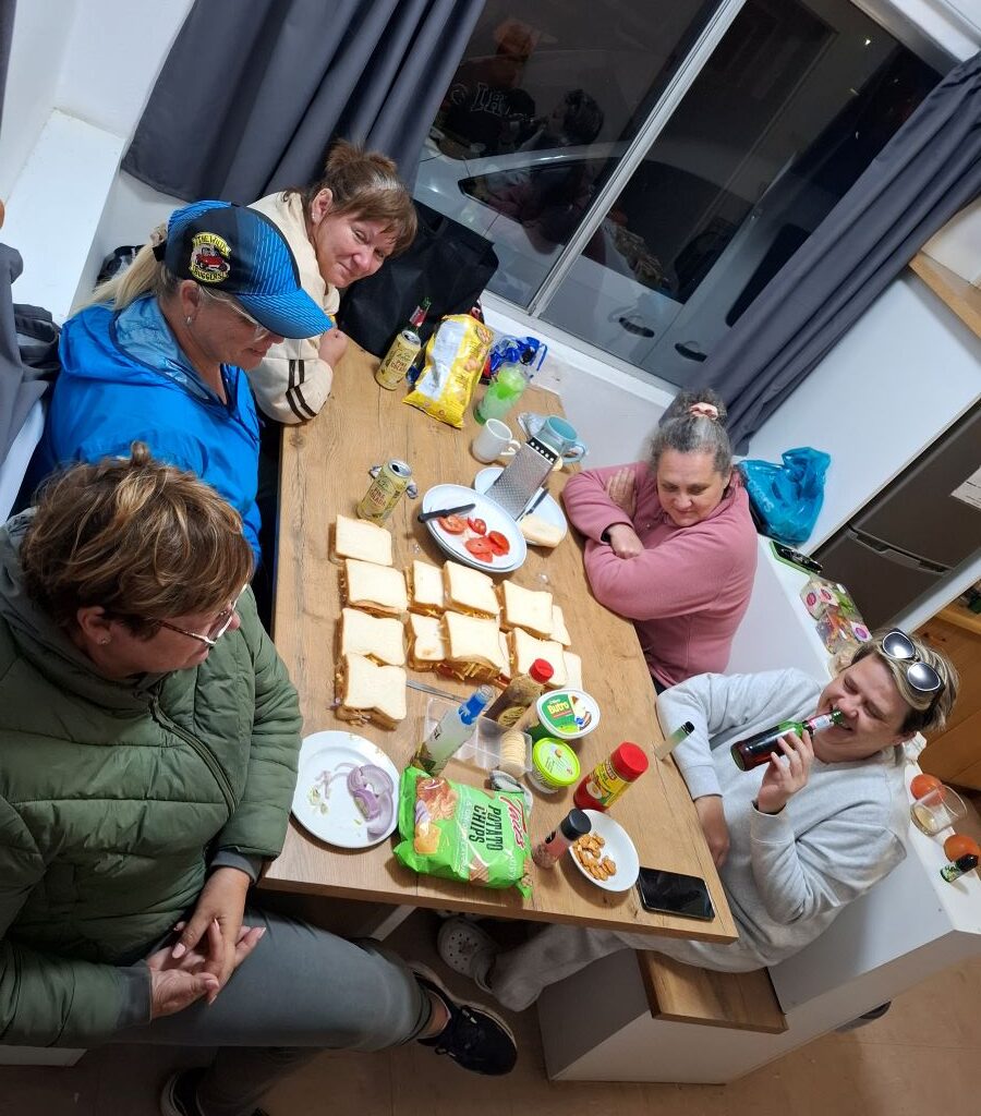 A warm, candid photo taken at the Bergriver Resort camp campsite after sunset, showing the Wild Buggers preparing for the braai, with the "Ladybugs" proudly displaying a tray of perfectly prepared braaibroodjies (toasted sandwiches).