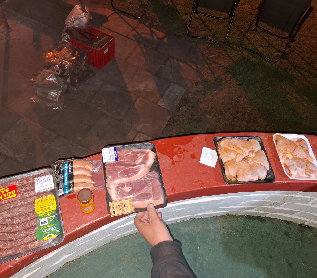 A visually appetizing photo of various cuts of marinated meat (steaks, chops, boerewors) neatly laid out on a table or counter, ready to be transferred to the roosters (braai grids) for the evening braai at the Bergriver Resort camp.