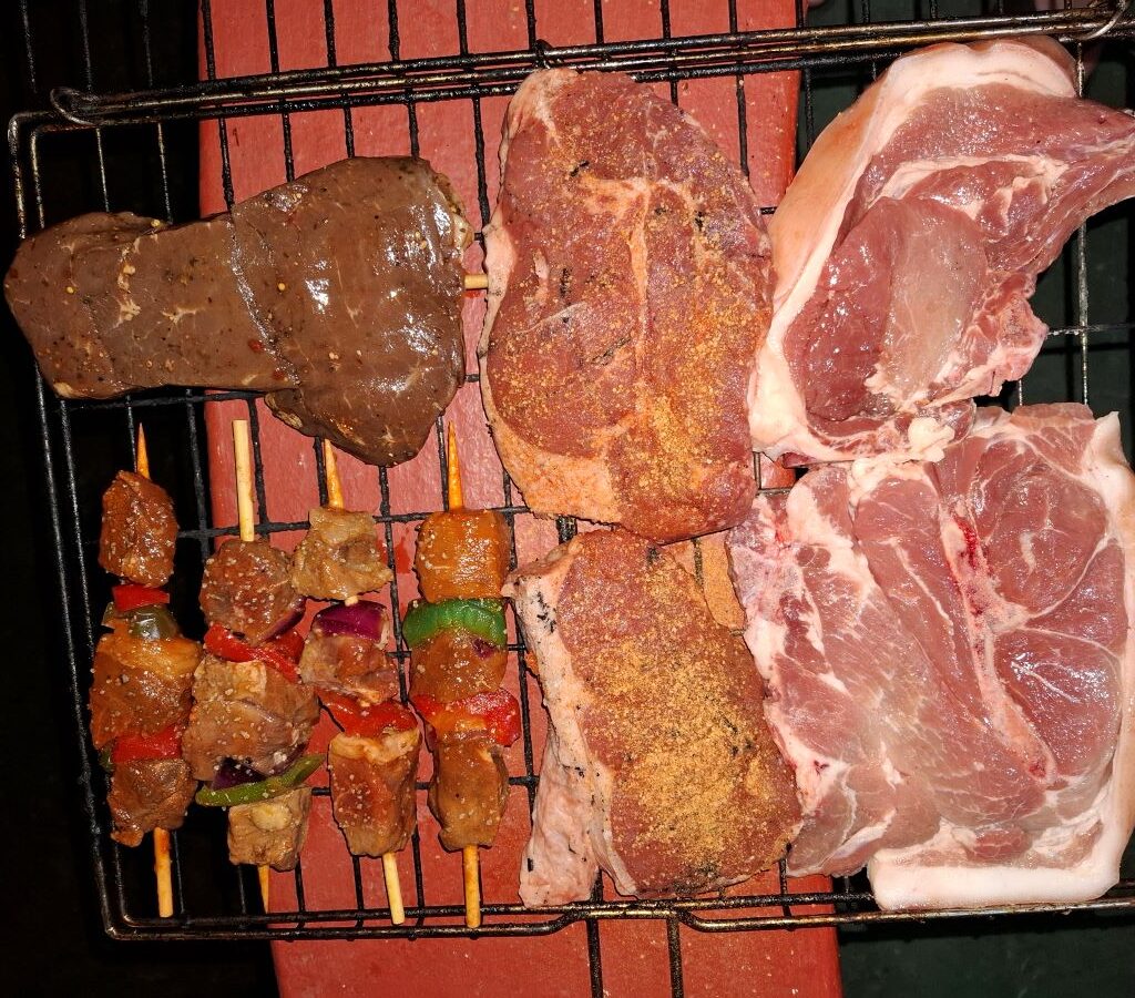 A close-up, mouth-watering photo of a braai rooster (grid) packed tightly with a magnificent assortment of meat, including thick pork chops, juicy steaks, savory sosaties (kebabs), and lamb, all ready for the Wild Buggers evening braai.