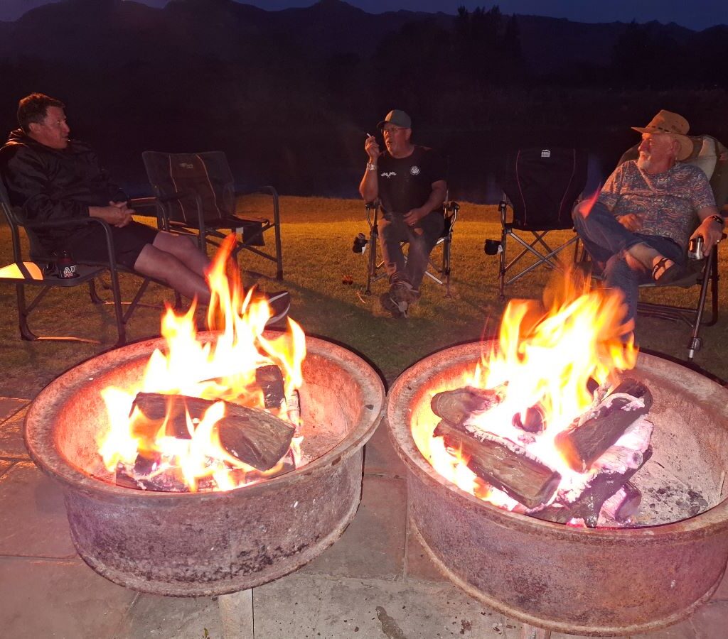 A dark, atmospheric photo taken late at night at the Bergriver Resort camp. The image shows three silhouettes—Anthony, Brent, and Jacques—still sitting closely around the glowing embers of a campfire, demonstrating the fun and extended camaraderie of the Wild Buggers club.