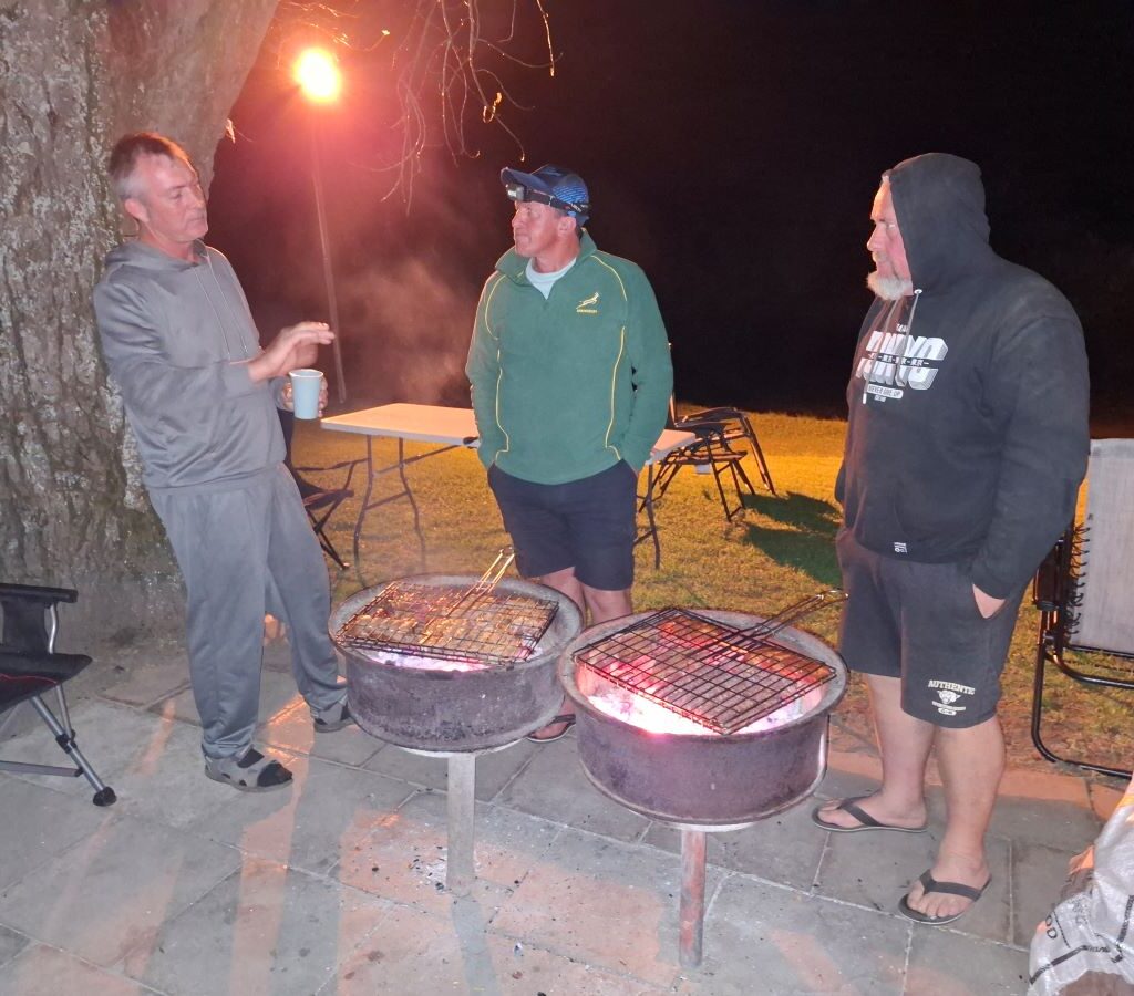 A warm, atmospheric photo of the Wild Buggers braai masters—Rhoodie, Brent, and Anton Horne—standing intently by the fire, monitoring the meat on the rooster (braai grid) under the dark evening sky at the Bergriver Resort camp.