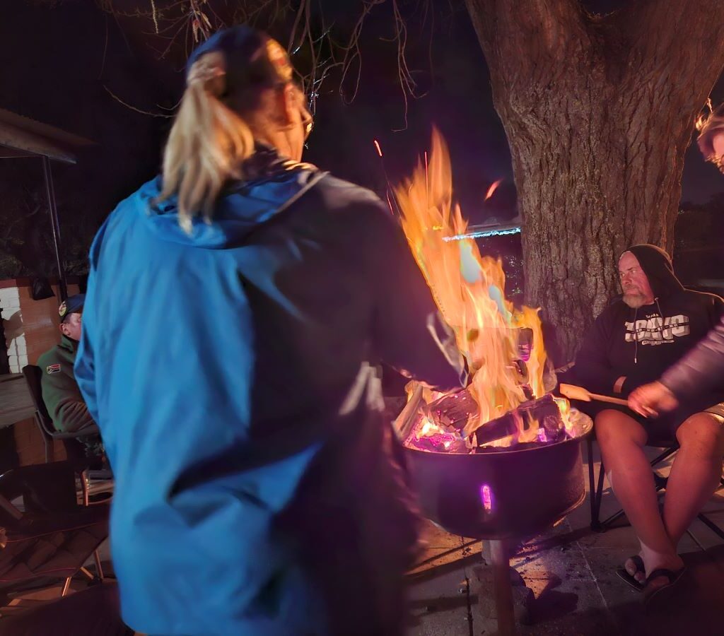 A warm, charming photo taken at the Bergriver Resort camp campsite showing Michelle and Coralie standing by the glowing campfire, happily toasting marshmallows on sticks, adding a cozy, sweet touch to the evening.