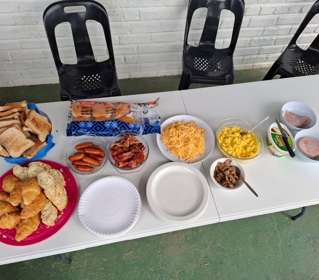 A wide, descriptive photo of the fully set breakfast table at the Wild Buggers Bergriver Resort camp campsite, showing plates, cutlery, and cups ready for use, capturing the quiet anticipation of the morning meal.