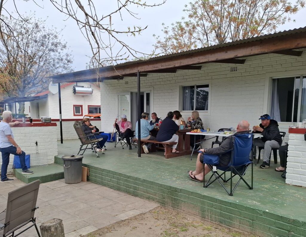 A lively photo of the Wild Buggers members gathered around the breakfast table at the Bergriver Resort camp campsite, cheerfully serving themselves a hearty meal, with coffee mugs in hand, signaling the start of a new day.