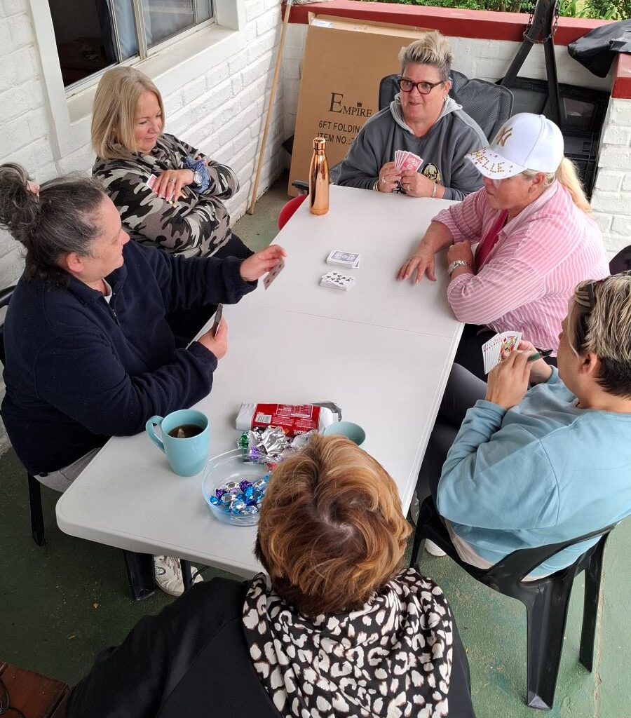 A joyful and humorous photo of several Wild Buggers "Ladybugs" (women members) sitting together and enthusiastically playing a game of cards, roaring with laughter during a relaxing moment at the Bergriver Resort camp.