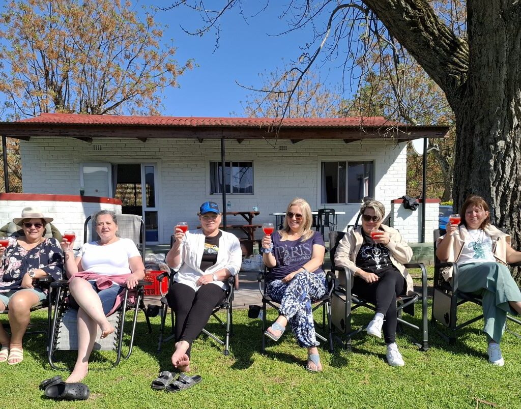 A joyful and sunny photo of six women from the Wild Buggers Beach Buggy Club, referred to as "Ladybugs," relaxing in camping chairs, posing with cocktails in hand and enjoying the sun at the Bergriver Resort camp. The image radiates fun and camaraderie.