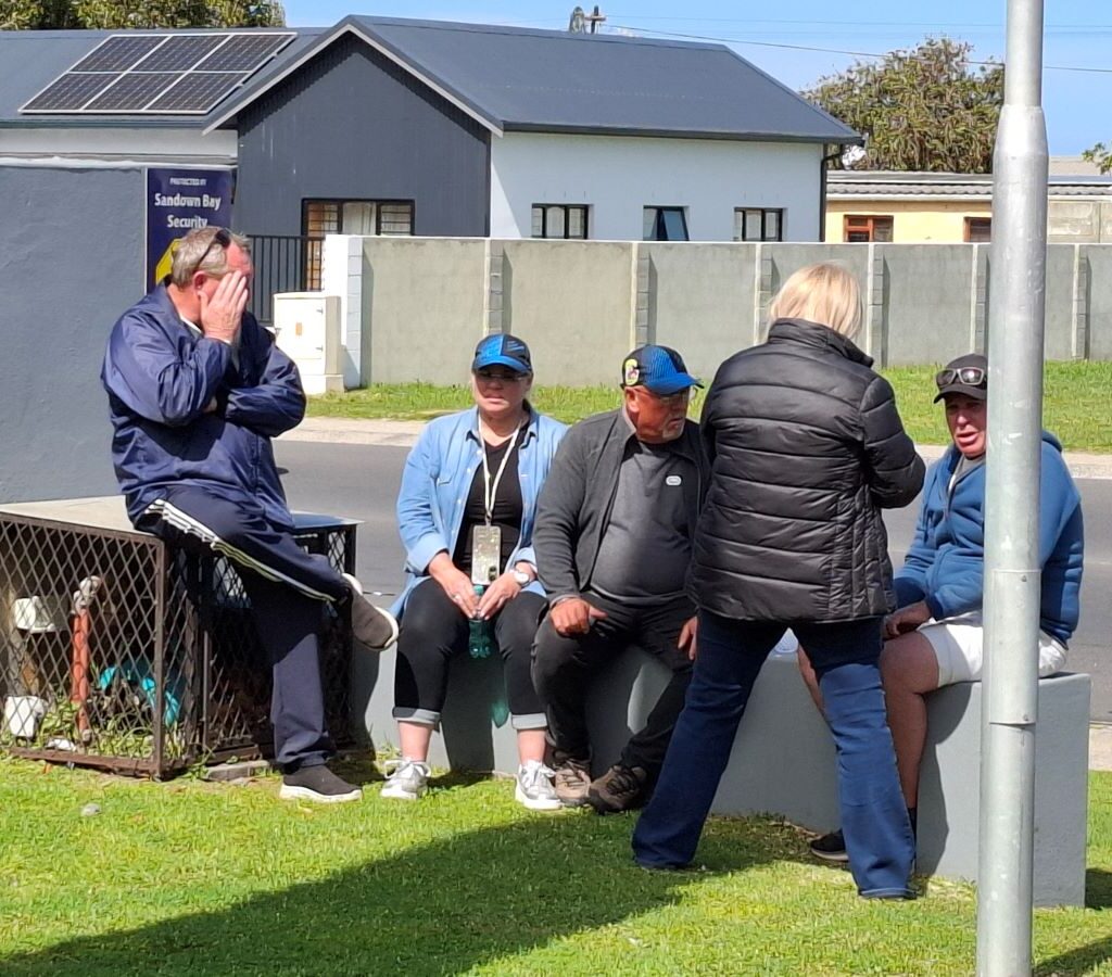 Several Wild Buggers members are pictured sitting on a wall at a garage in Kleinmond, chatting during a road trip stop.