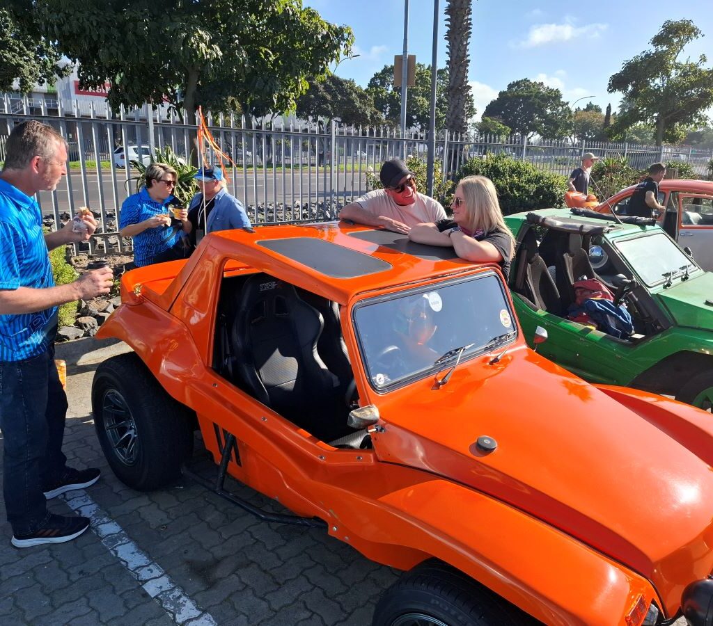 A group of Wild Buggers chat and drink coffee around Brent's orange VW air-cooled Salamander beach buggy.