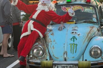 A person dressed as Santa Claus posing on the hood of Russell's brightly decorated VW Beetle at the Annual Lights For Life Run meet-up.