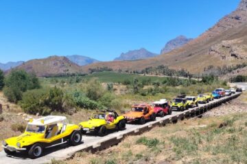 A long line of colorful beach buggies, a red and white VW Kombi, and a white Baja Bug crossing a narrow single-lane concrete bridge over a river.