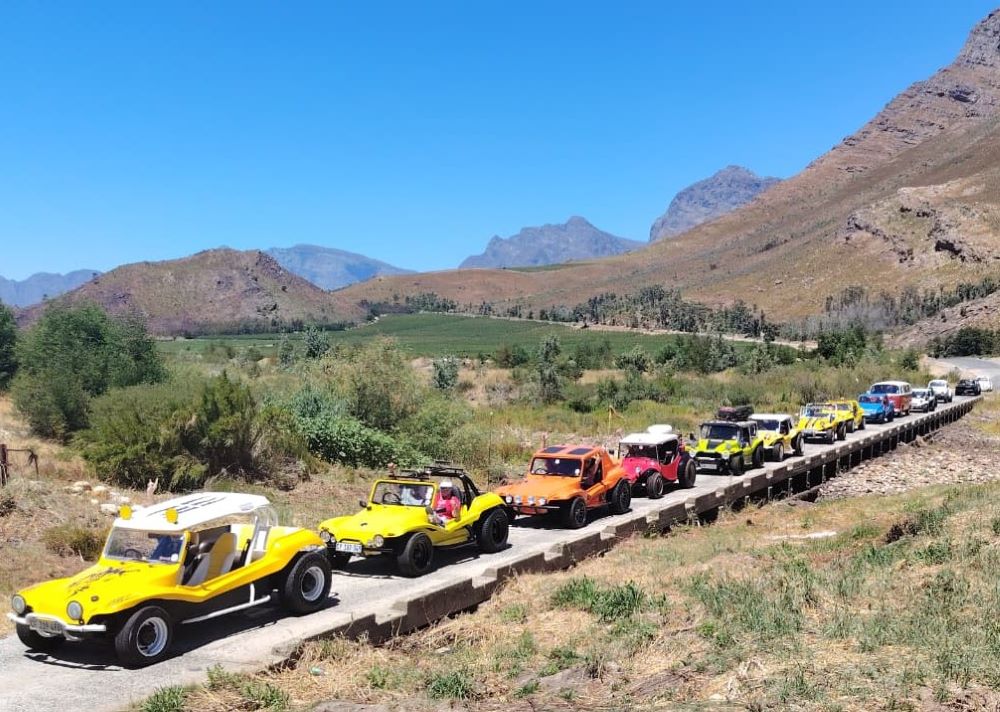A long line of colorful beach buggies, a red and white VW Kombi, and a white Baja Bug crossing a narrow single-lane concrete bridge over a river.