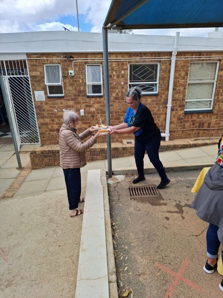 Mickey handing two dished-up plates of food to Jo-ann Gericke to serve to the residents at Goue Aar.