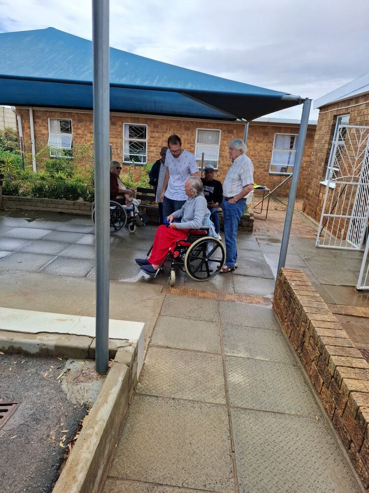 A group of residents and staff members, one in a wheelchair, gathered outside the Goue Aar Old Age Home facility after a Wild Buggers Beach Buggy Club visit.