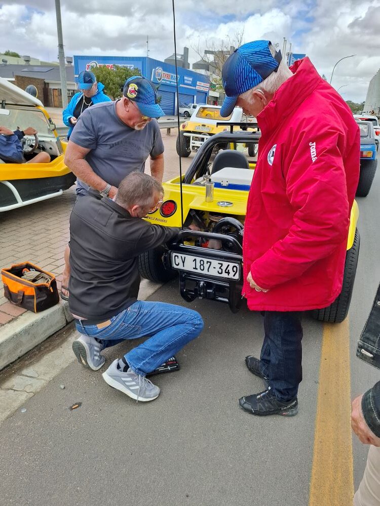 Three Wild Buggers Beach Buggy Club members huddled behind a yellow beach buggy, inspecting the air-cooled VW engine on the side of the road.