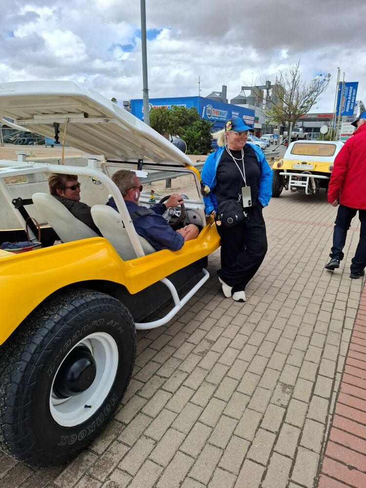 A group of The Wild Buggers Beach Buggy Club members waiting on a brick-paved sidewalk in Malmesbury while a yellow beach buggy is being repaired.