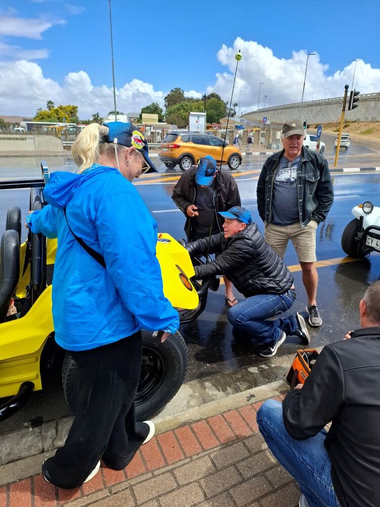 Wild Buggers club members wearing blue branded caps repairing a yellow beach buggy on a wet roadside during a rain shower.