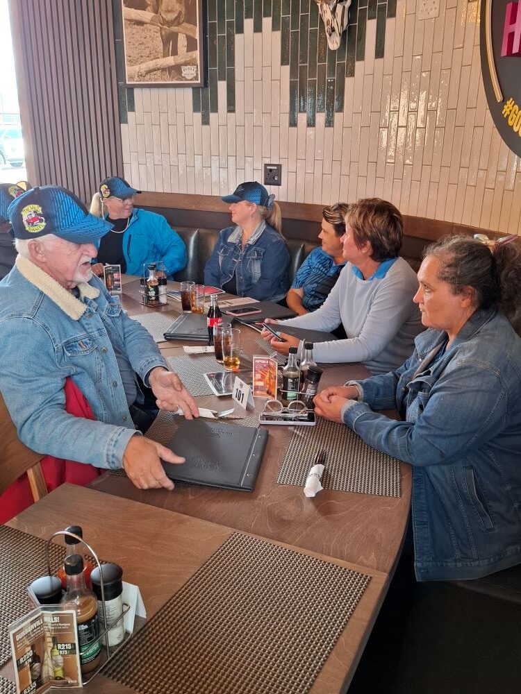 Members of The Wild Buggers Beach Buggy Club wearing official blue and black branded club caps while enjoying a meal together.