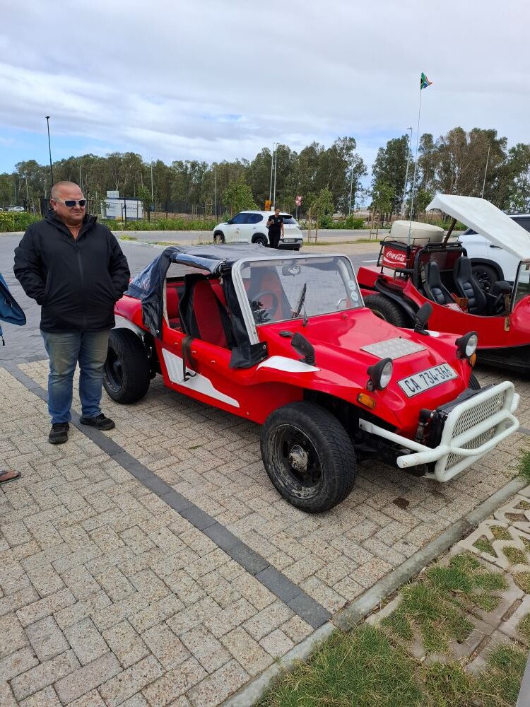 Ivor Du Plessis standing next to his custom red Beach Buggy, which features a prominent white front radiator guard and red-and-white accents.