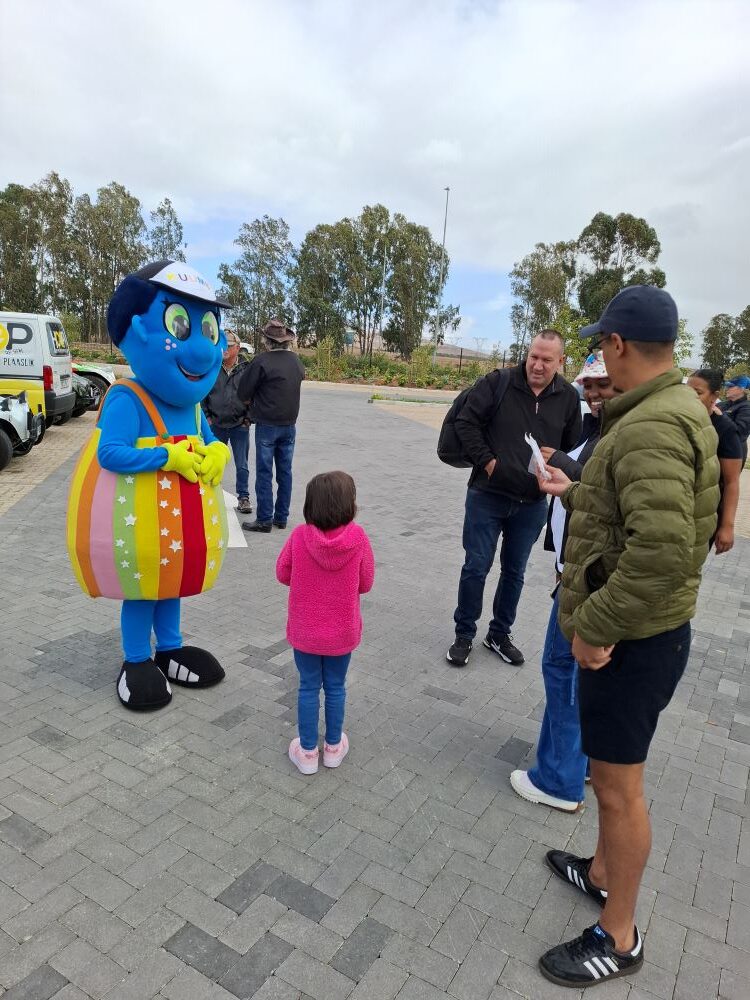Alt Text: The Badisa charity mascot, a blue character in a rainbow-striped outfit, meeting a child and Wild Buggers club members at the Groot Phesantekraal meeting point.