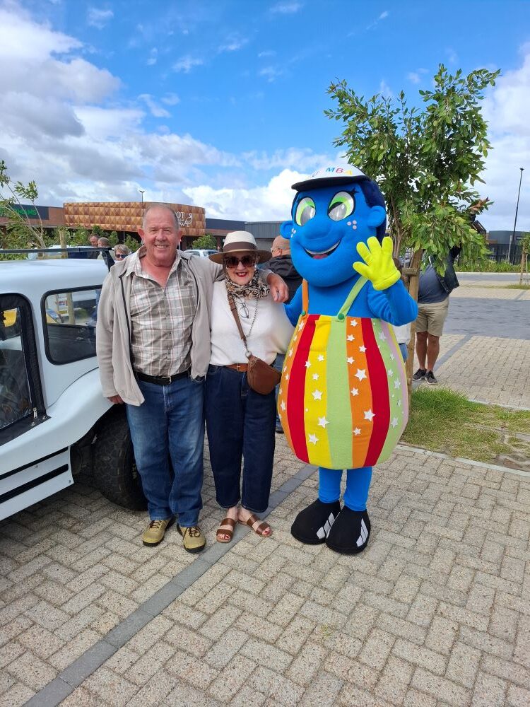 Andre and Jo-Ann Gericke from The Wild Buggers Beach Buggy Club pose with the blue and rainbow-striped Badisa charity mascot next to their white beach buggy.