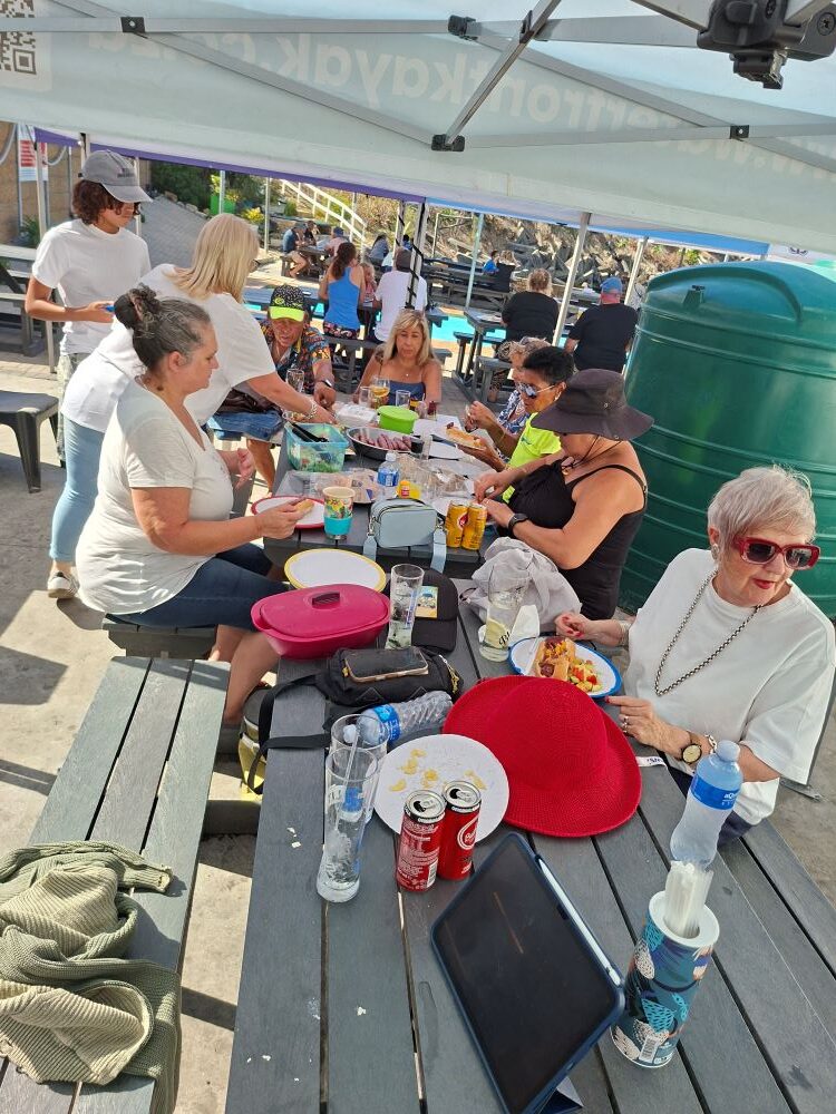 A group of Wild Buggers Beach Buggy Club members sitting at outdoor picnic tables under a gazebo, enjoying a braai meal at the Oceana Power Boat Club.