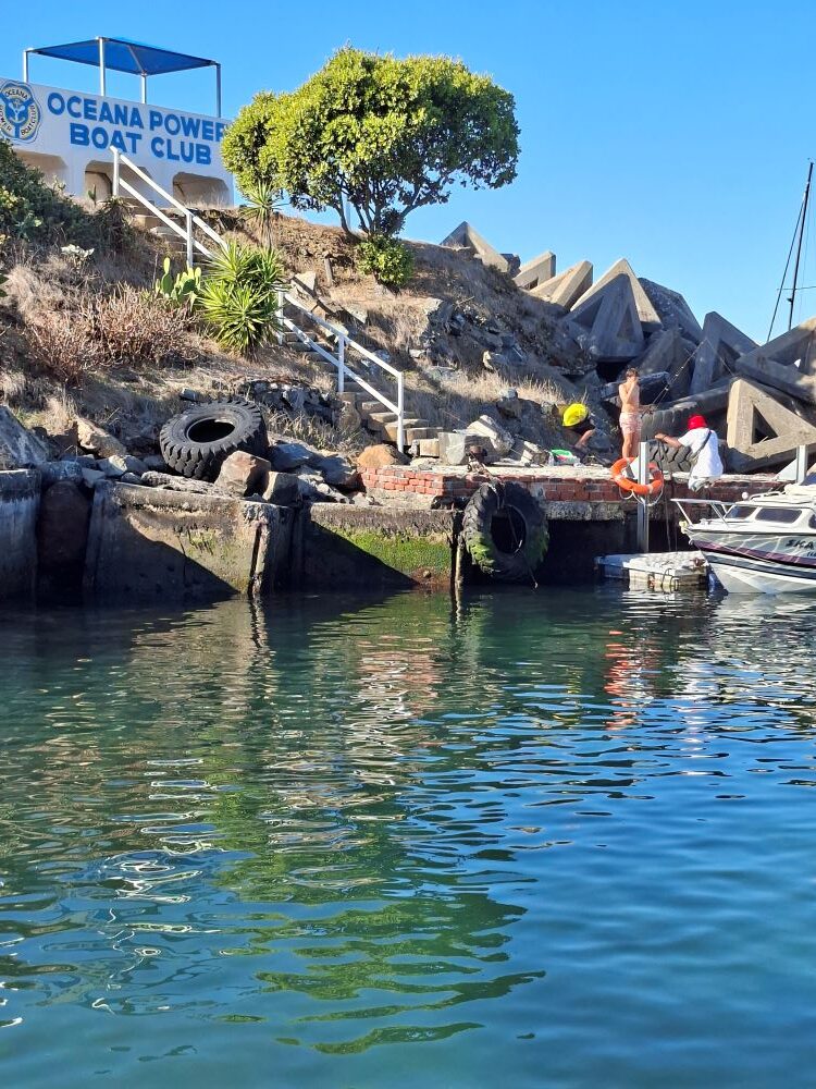 A scenic view of the Oceana Power Boat Club clubhouse and harbor, featuring rugged rocks, green foliage, and the calm Atlantic ocean water in the foreground.