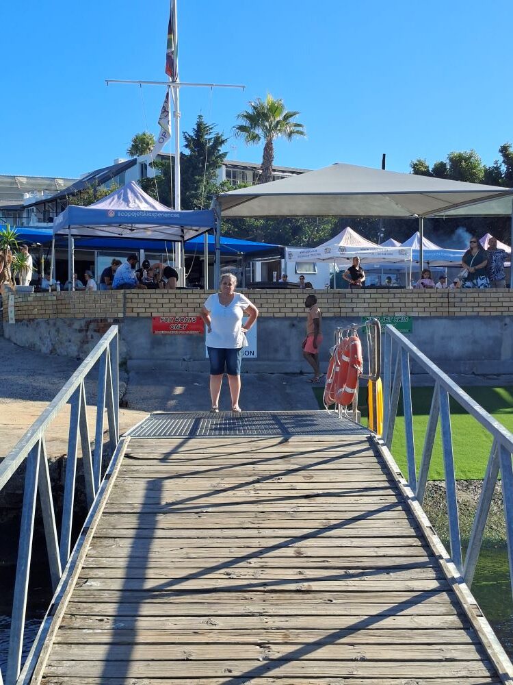 A member of the Wild Buggers Beach Buggy Club standing on a wooden jetty bridge at the Oceana Power Boat Club with the clubhouse and Atlantic ocean in the background.