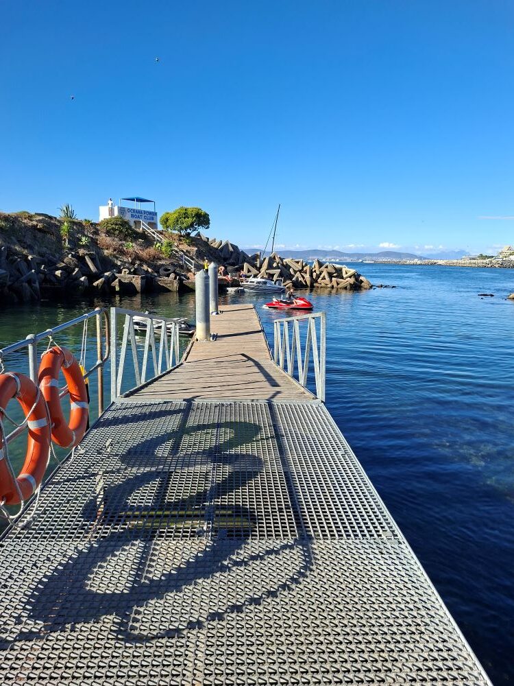 A wide scenic view from a metal mesh jetty at the Oceana Power Boat Club showing the blue Atlantic water, the clubhouse on the rocks, and a red jet ski in the harbor.