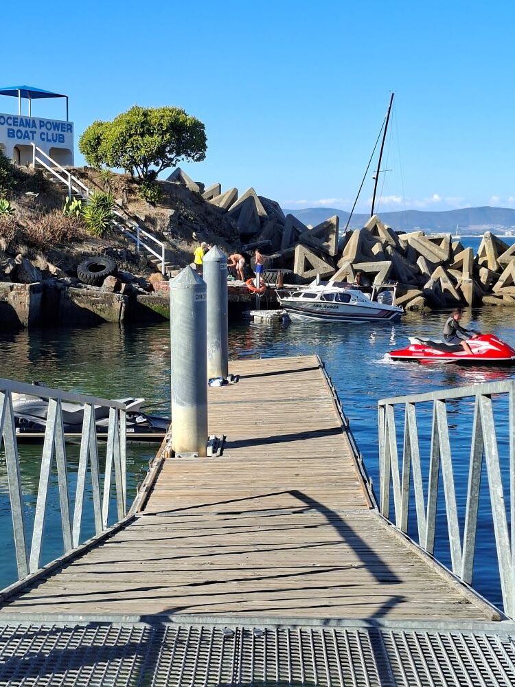 A view from a wooden jetty at the Oceana Power Boat Club showing a motorboat docked and a red jet ski moving through the water near the rocky harbor shore.