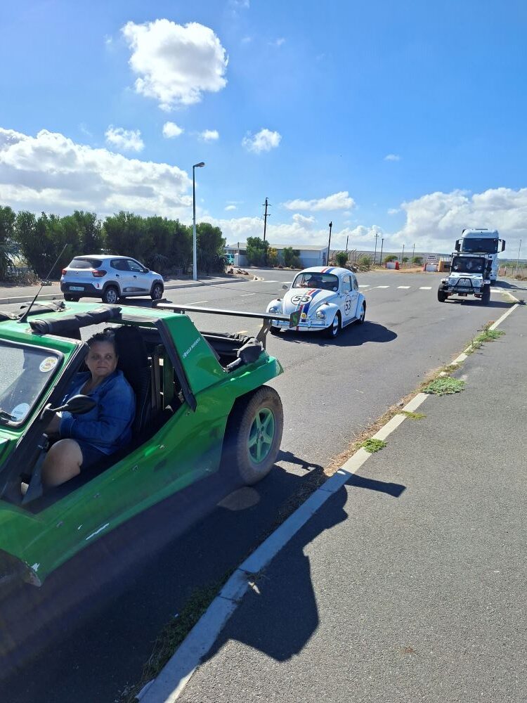 A convoy of Wild Buggers vehicles including a green Kango beach buggy, an iconic Herbie VW Beetle, and a white Jeep driving toward the Waterfront in Cape Town.
