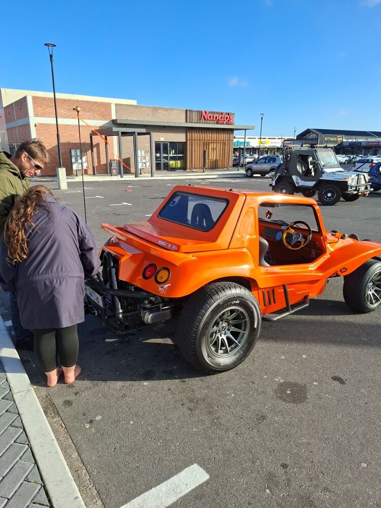 Lerene and her partner standing behind Brent and Sam Van Der Weijde’s orange Salamander beach buggy, The Wasp, at the Bothasig meeting point while inspecting the 1600 VW air-cooled motor before the Wild Buggers West Coast cultural experience Run.