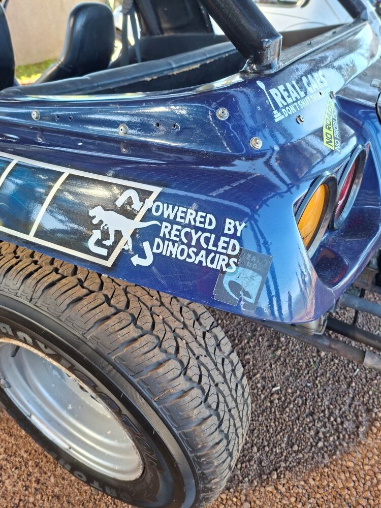 A close-up of a humorous bumper sticker reading "powered by recycled dinosaurs" on Lerene's custom beach buggy during the Wild Buggers West Coast cultural experience Run convoy.