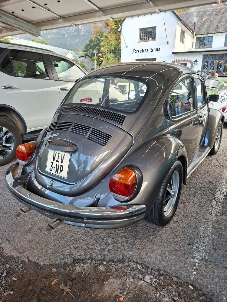 A three-quarter rear view of Viv Reyneke’s pristine metallic gunmetal grey classic VW Beetle 1600 with custom chrome bumpers, dual exhaust tips, and personalized Western Cape license plates, parked at the Foresters Arms meeting point.