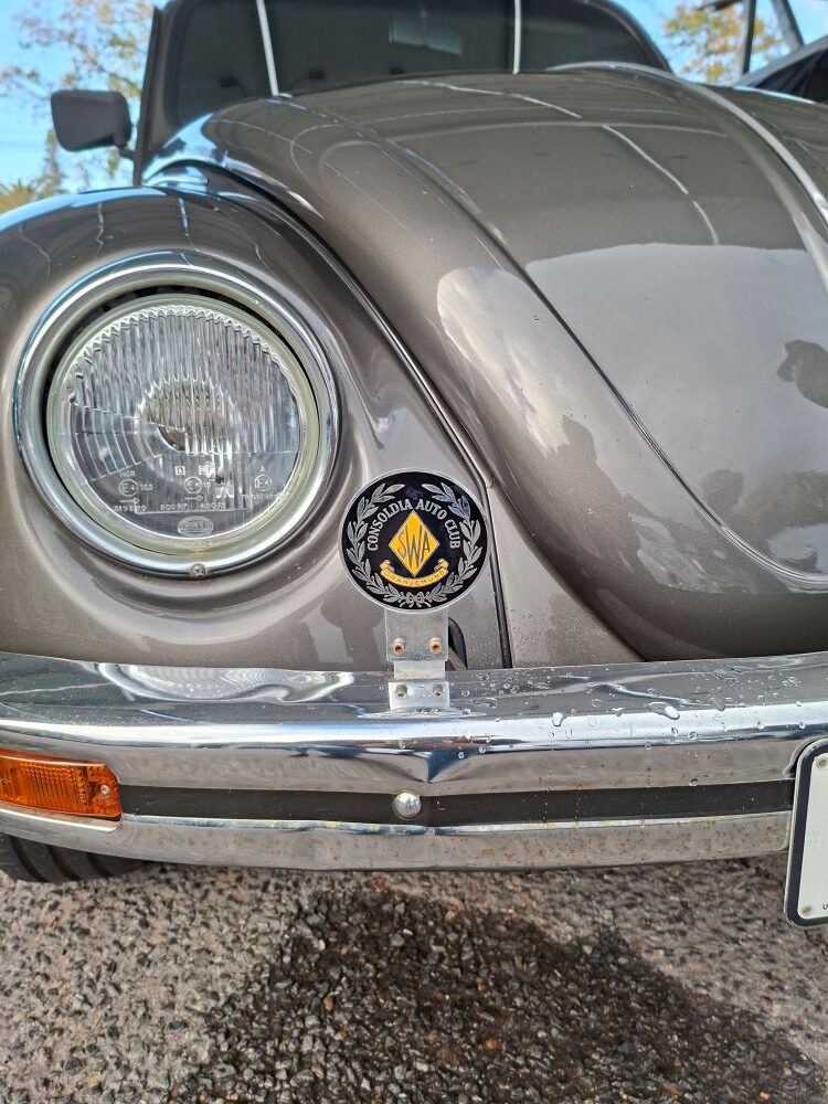 A close-up front view of Viv Reyneke’s metallic gunmetal grey classic VW Beetle, focusing on a vintage round Consoldia Auto Club Oranjemund SWA badge mounted to the chrome bumper next to a Hella headlight.