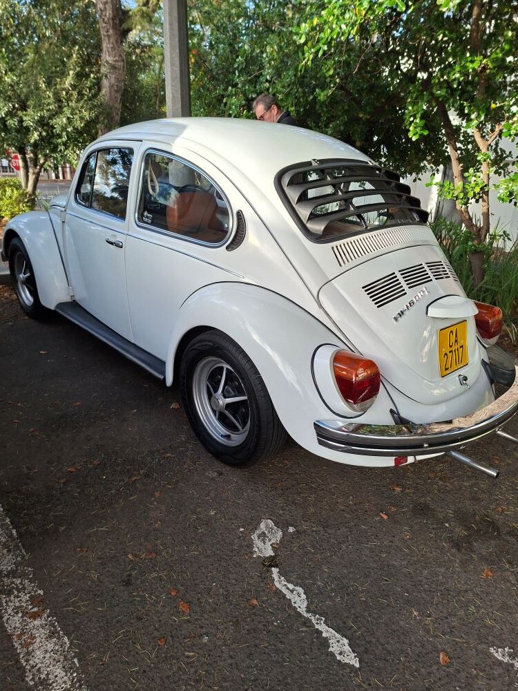 A clean three-quarter rear view of Andre and Jo-ann Gericke’s classic white 1975 VW Beetle 1600 featuring a black rear window louvre, custom alloy wheels, and a vintage yellow Cape Town license plate, parked under green trees at a car club event.